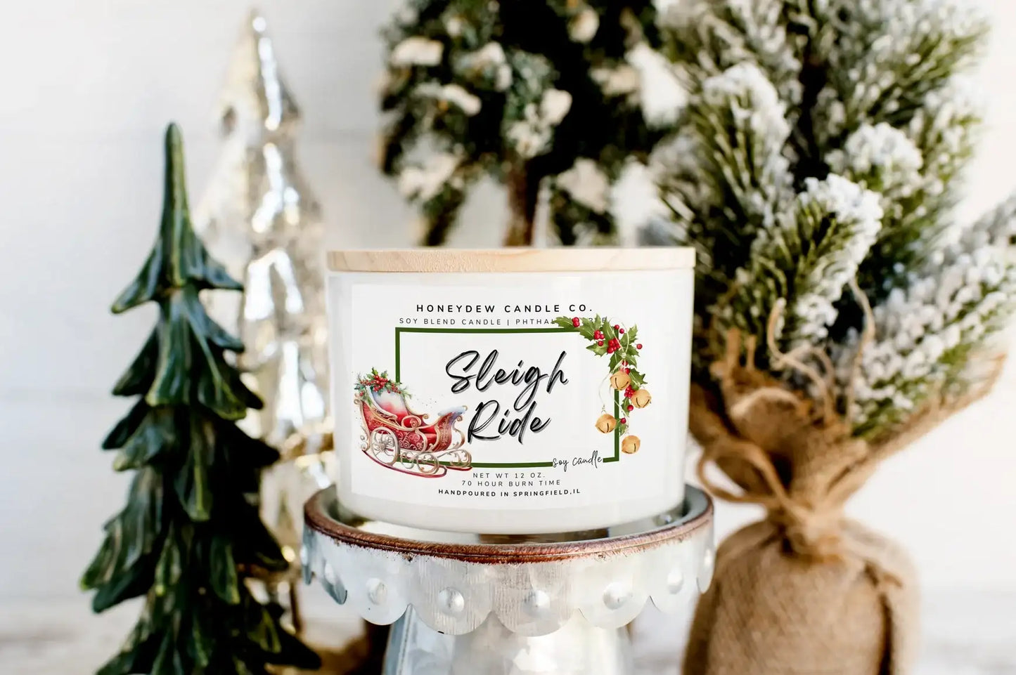 Candle with 'Sleigh Ride' label in front of decorative Christmas trees