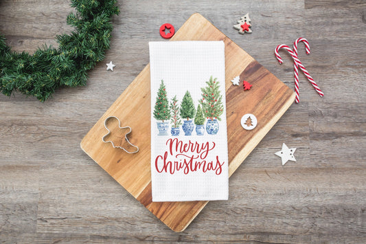 Christmas-themed kitchen towel with trees and 'Merry Christmas' text on a wooden board with festive decorations.