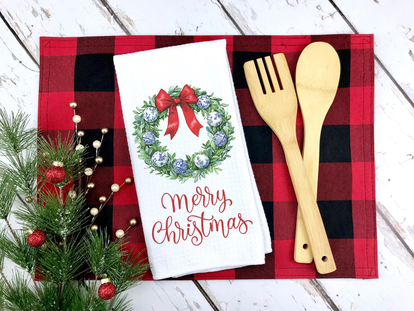 Christmas-themed towel with wreath and 'Merry Christmas' text on a red and black checkered tablecloth with wooden utensils.