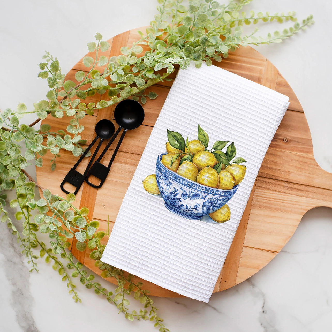 White towel with a lemon design on a wooden cutting board with black spoons and greenery.