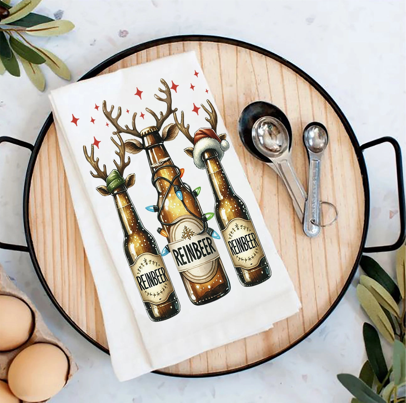 Tea towel with reindeer-themed beer bottles on a wooden board with measuring spoons and eggs.