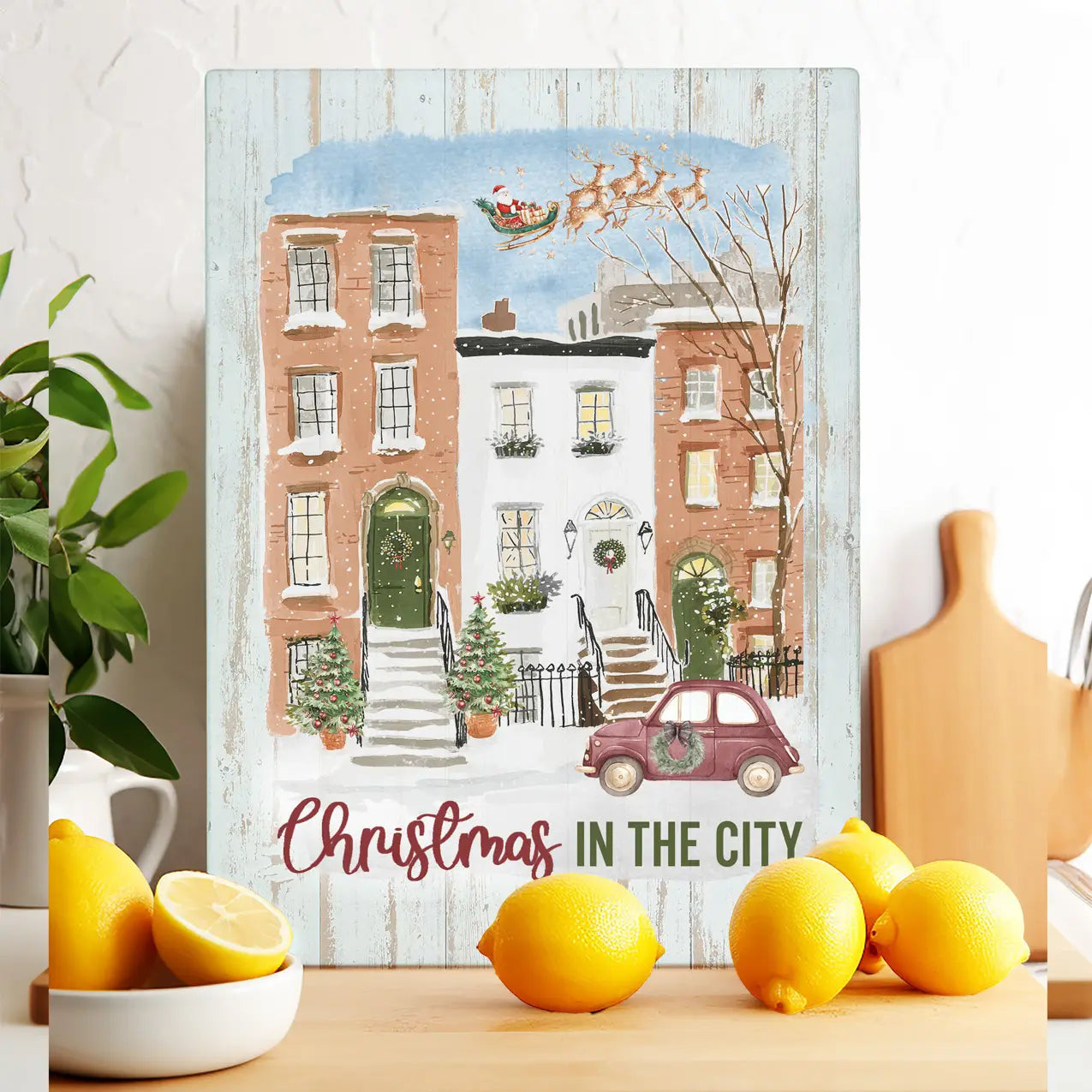 A rectangular decorative sign or print featuring a whimsical winter city street scene with snow-covered brick and white buildings, a red car, a Santa sleigh, and the text 'Christmas in the City,' styled on a tabletop with lemons and kitchen props.