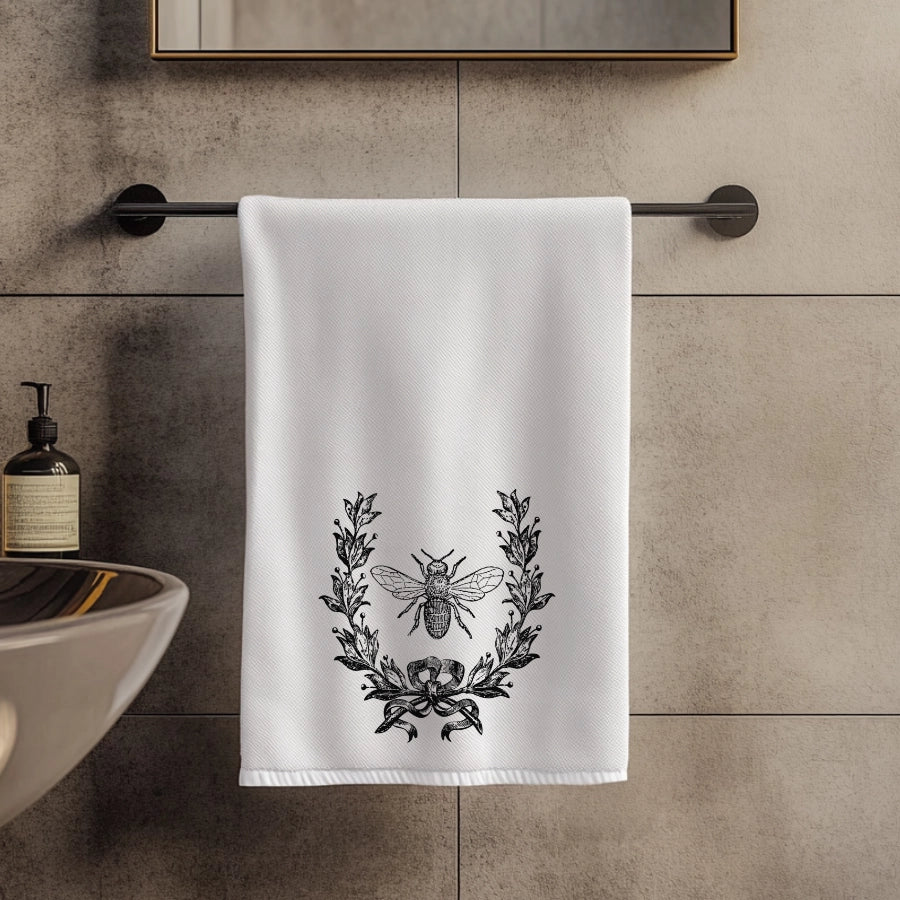 White towel with black laurel wreath and bee design hanging on a towel rack in a bathroom.