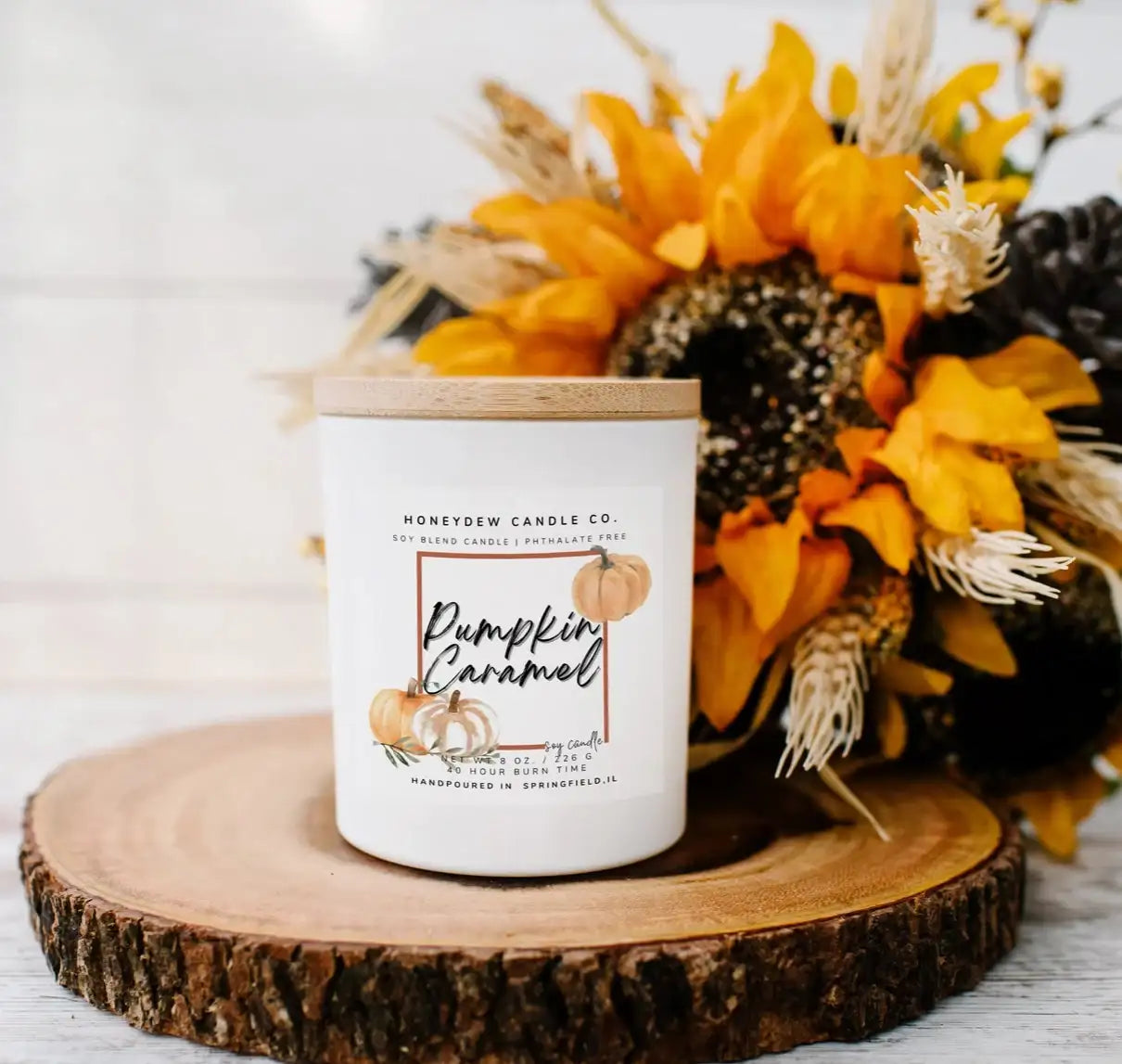 Candle labeled 'Pumpkin Caramel' on a wooden coaster with sunflowers and pinecones.
