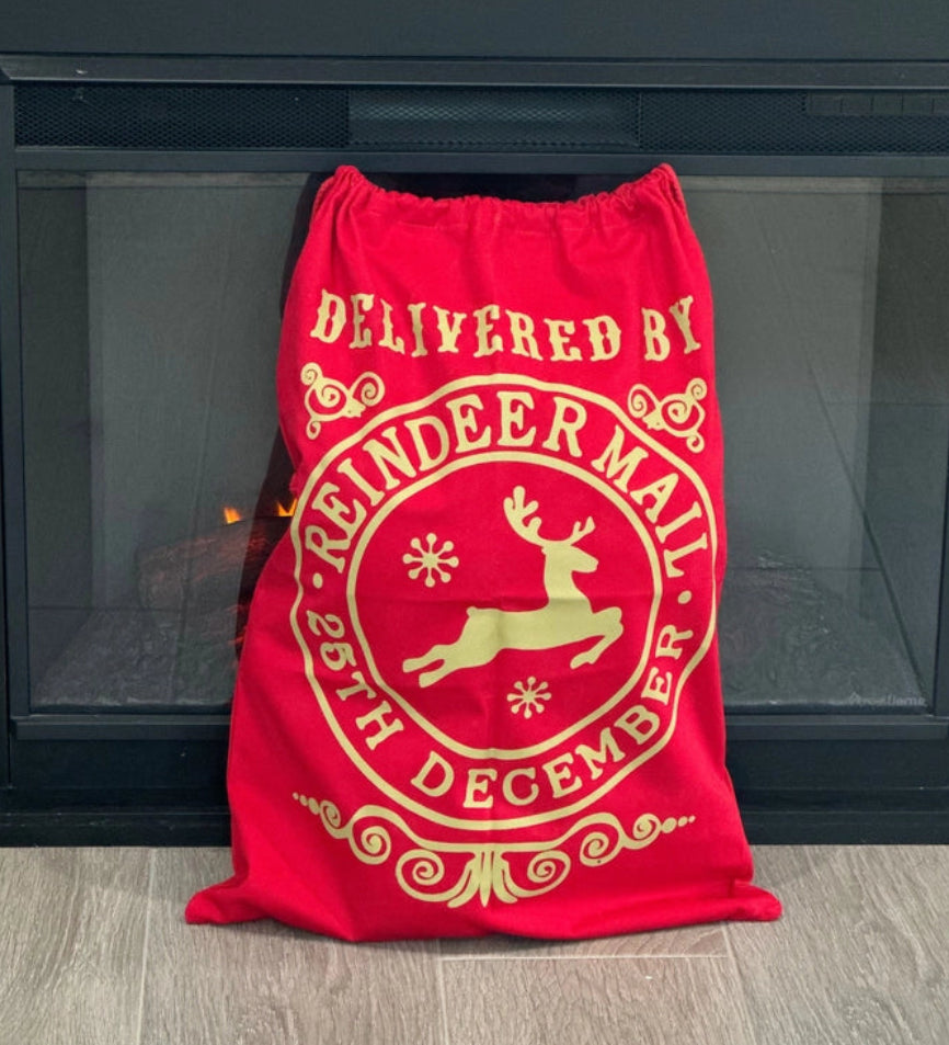 Red cotton Reindeer Mail Christmas Santa Sack with gold reindeer design and drawstring closure, displayed in front of a fireplace.
