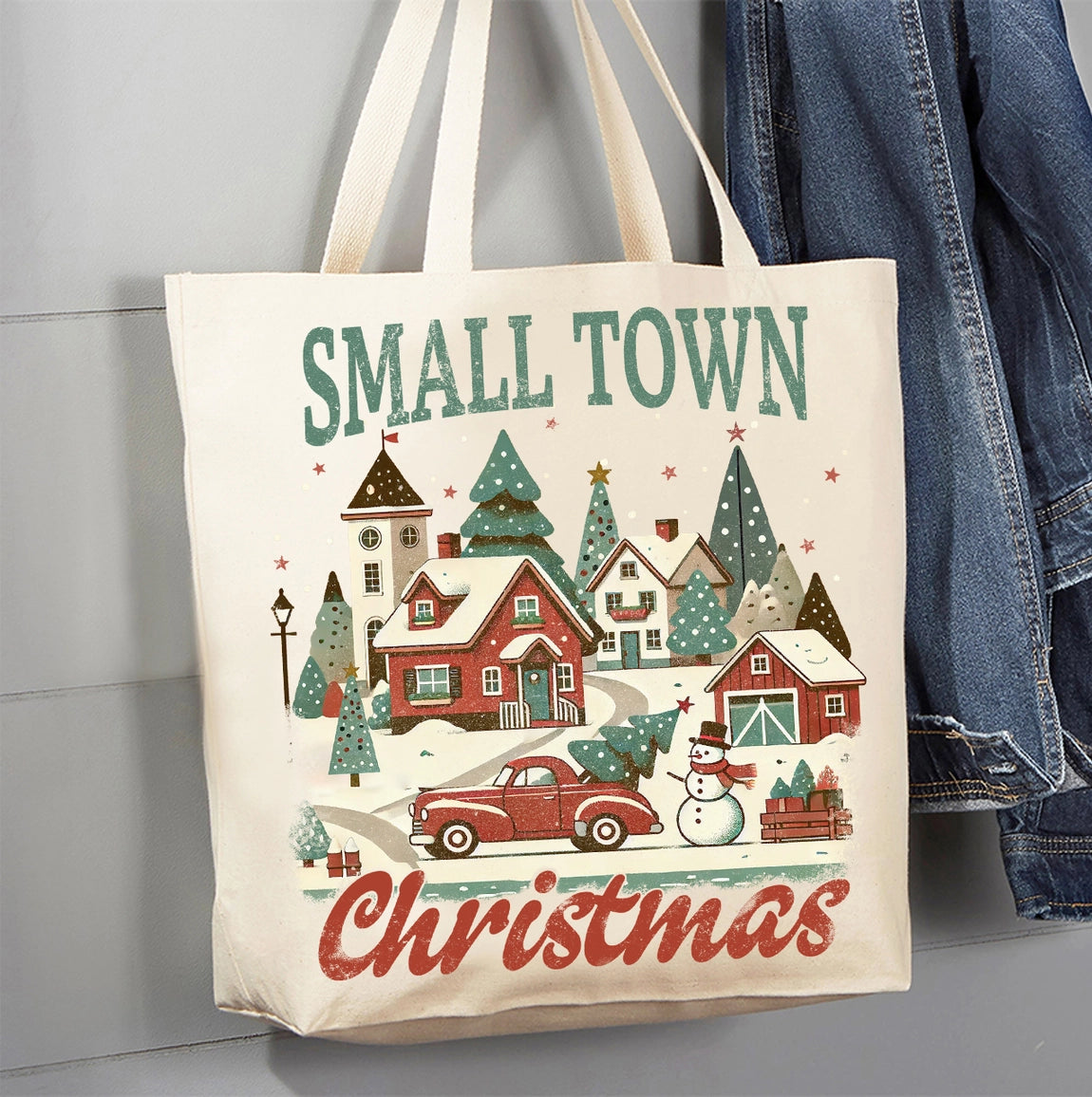 A cream canvas tote bag with a festive Christmas winter village print, featuring the words 'SMALL TOWN Christmas' in green and red lettering. The bag is hanging next to a pair of blue denim jeans against a gray paneled wall.