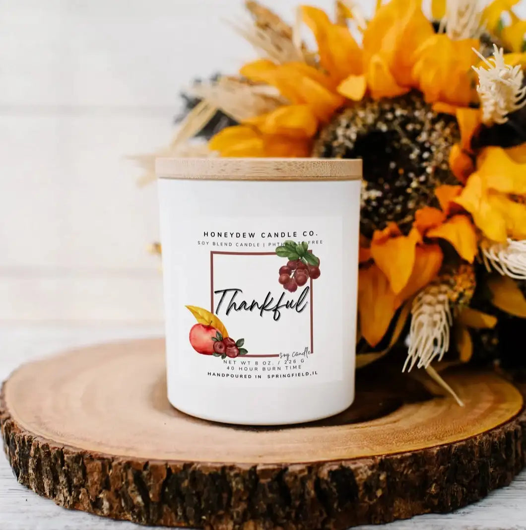 Candle labeled 'Thankful' on a wooden surface with sunflowers and pinecones.