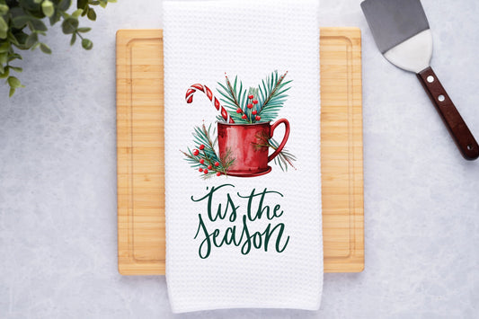Towel with Christmas design and 'tis the season text on a cutting board.