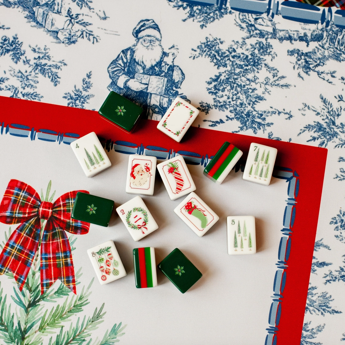 Christmas Mahjong tiles displayed on Tartan Toile Noël mat featuring red tartan bow, blue-and-white toile and holiday greenery.