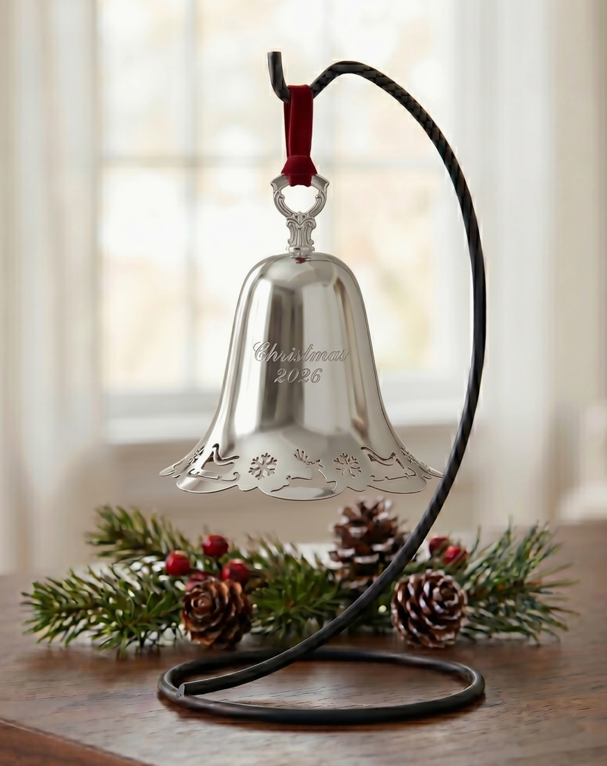 2026 Towle silver-plated Christmas bell ornament displayed on a stand with festive evergreen branches, berries and pinecones.