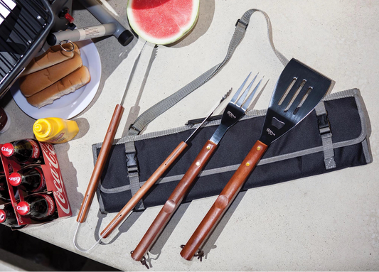 Barbecue tools with a grill, hot dogs, buns, and watermelon on a table.