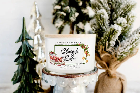 Candle with 'Sleigh Ride' label in front of decorative Christmas trees