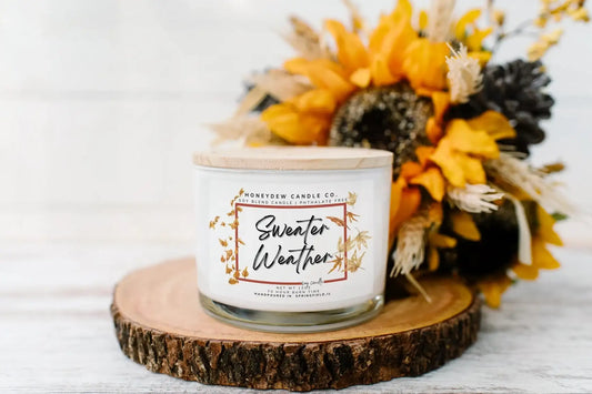 Candle labeled 'Sweater Weather' on a wooden coaster with sunflowers and pinecones.