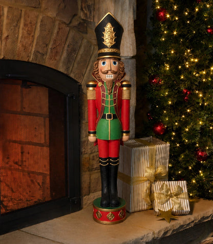 A large wooden nutcracker figurine stands on a stone fireplace mantel beside a lit fireplace, with wrapped gifts and a decorated Christmas tree in the background.