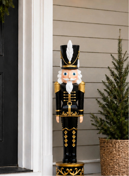 A large black and gold Christmas nutcracker decoration stands on a porch beside a black door, with a beige siding wall and a small potted tree in the background.