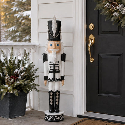 A large decorative nutcracker figurine stands on a front porch beside a black door and a potted evergreen arrangement. The nutcracker is dressed in a white and black uniform with a black hat featuring a white feather and silver trim, and a pale peach face and beard.