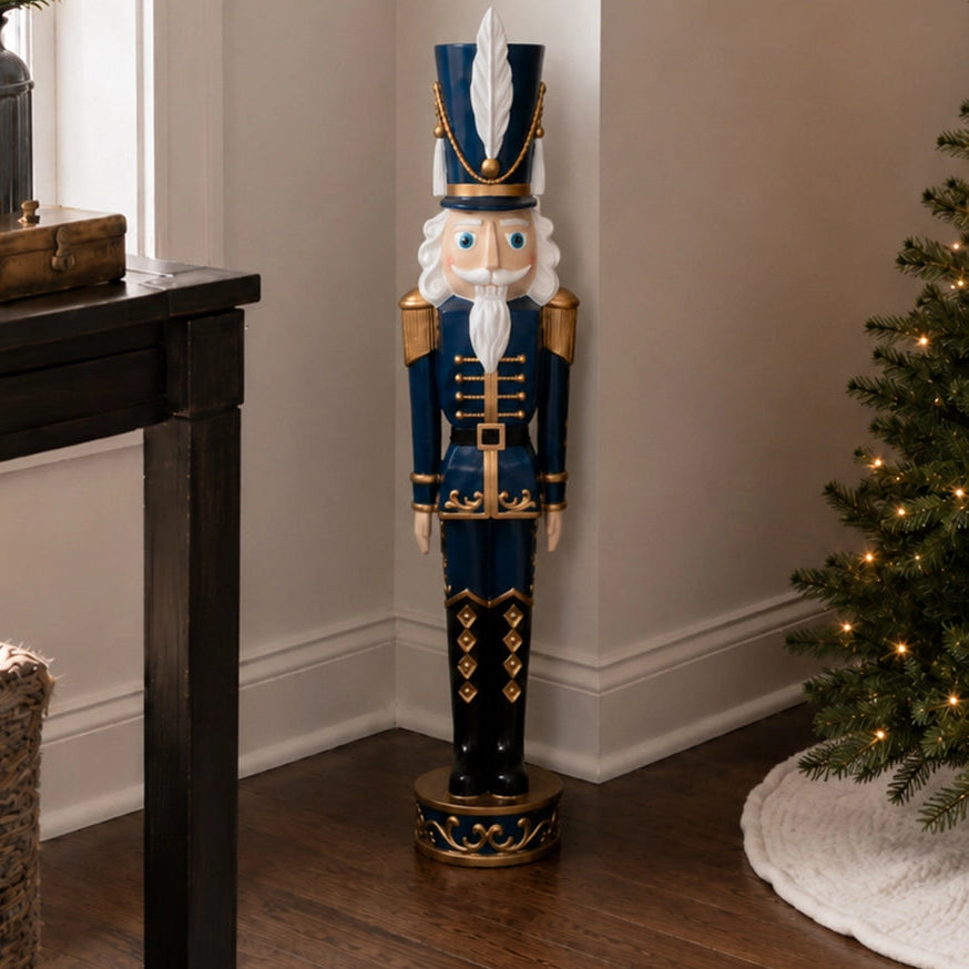 A tall decorative nutcracker figurine stands in a holiday room corner beside a black table and a lit Christmas tree. The figurine is dark blue with gold trim, a white beard, and a white feather on its hat.