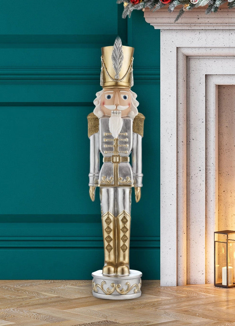 A large decorative nutcracker figurine stands on a wooden floor beside a fireplace. The nutcracker is metallic silver and gold with a white beard and face, and the scene includes a teal wall, white fireplace mantel, holiday garland, and lit lanterns.