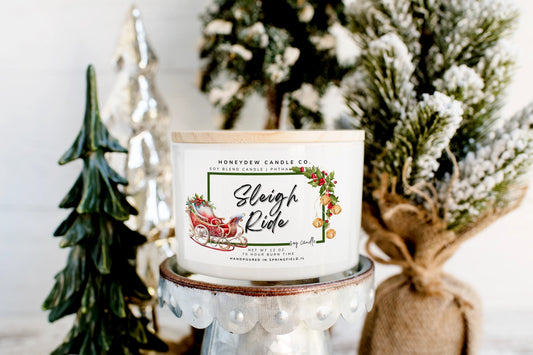 Candle with 'Sleigh Ride' label in front of decorative Christmas trees