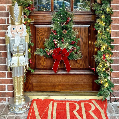 A festive holiday front door decorated with a green garland, a red bow, pinecones, and red berries, alongside a large silver and gold nutcracker and a red doormat with gold lettering.
