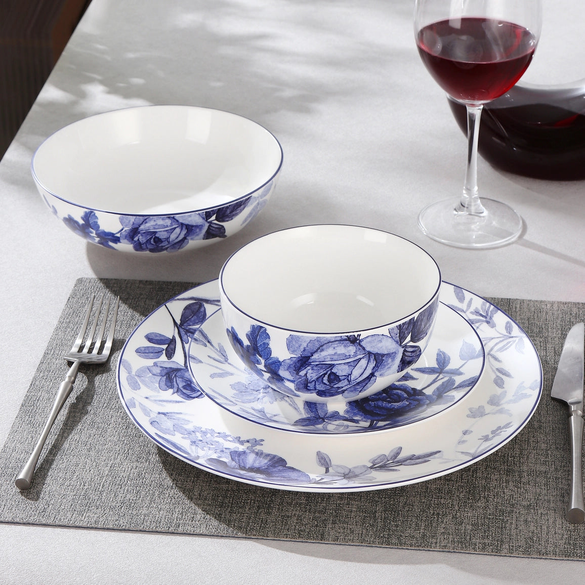 Blue and white floral ceramic dinnerware set on a dining table, including a plate, bowl, and matching cup, styled with silverware, a placemat, and a glass of red wine.