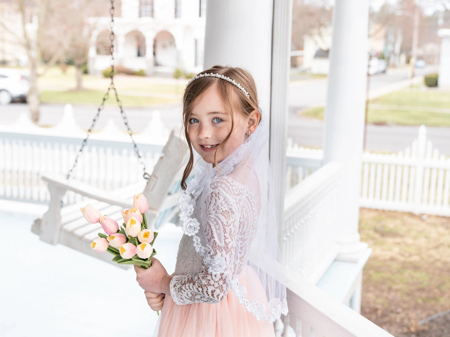 Young girl in a white lace dress holding flowers on a porch swing.
