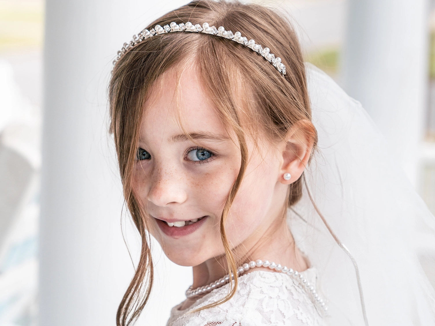 Young girl in a white dress with a headband and veil, smiling outdoors.