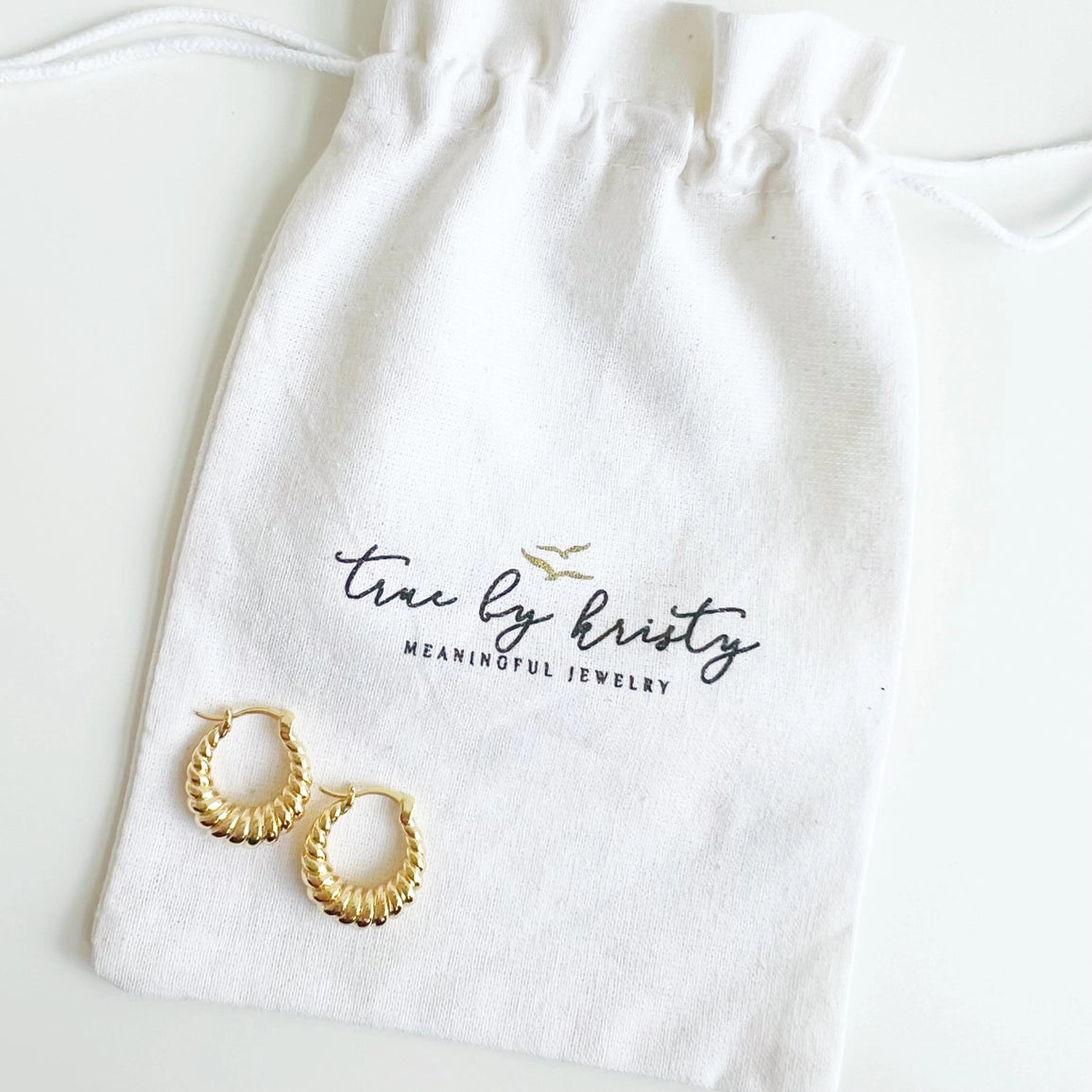 Gold hoop earrings on a white drawstring bag with 'true by kristy' branding.