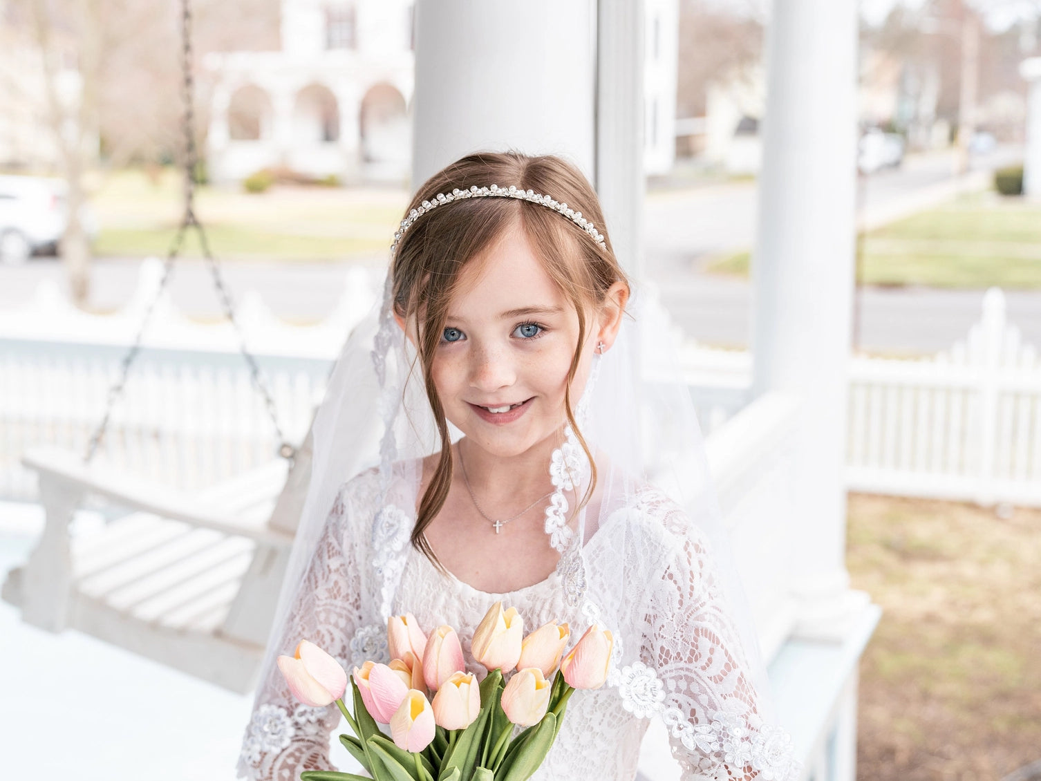 Young girl in a white dress with a veil holding tulips, standing on a porch.