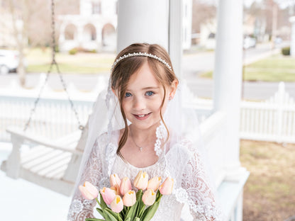 Young girl in a white dress with a veil holding tulips, standing on a porch.