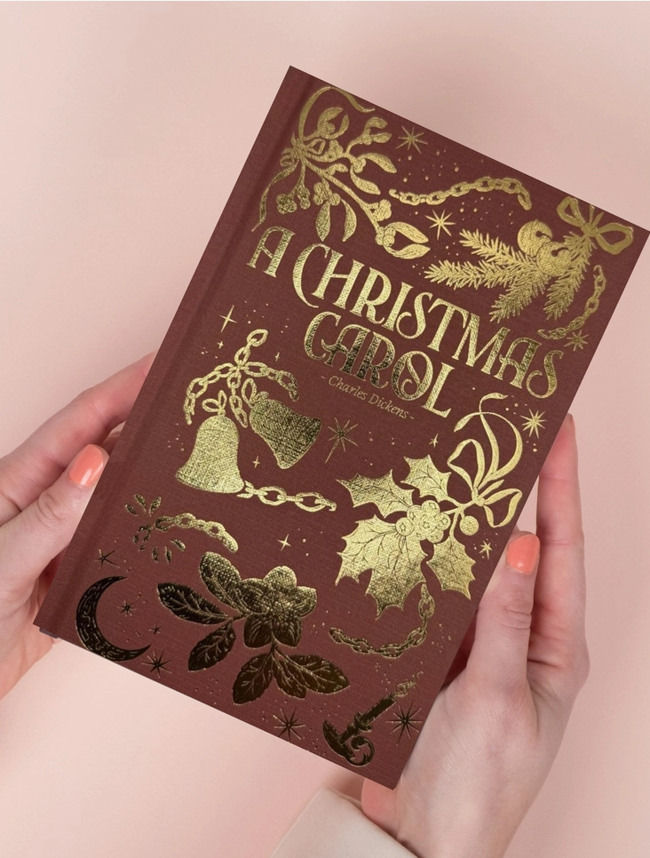 Book titled 'A Christmas Carol' with decorative cover held by hands against a light pink background