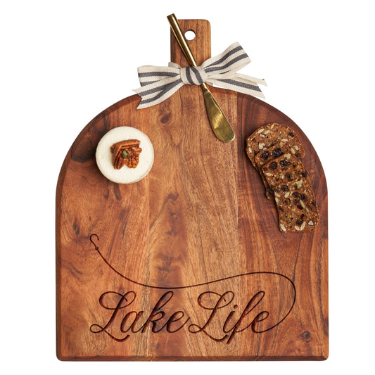Wooden cutting board with 'Lake Life' engraving, accompanied by a gold knife and food items on a white background.