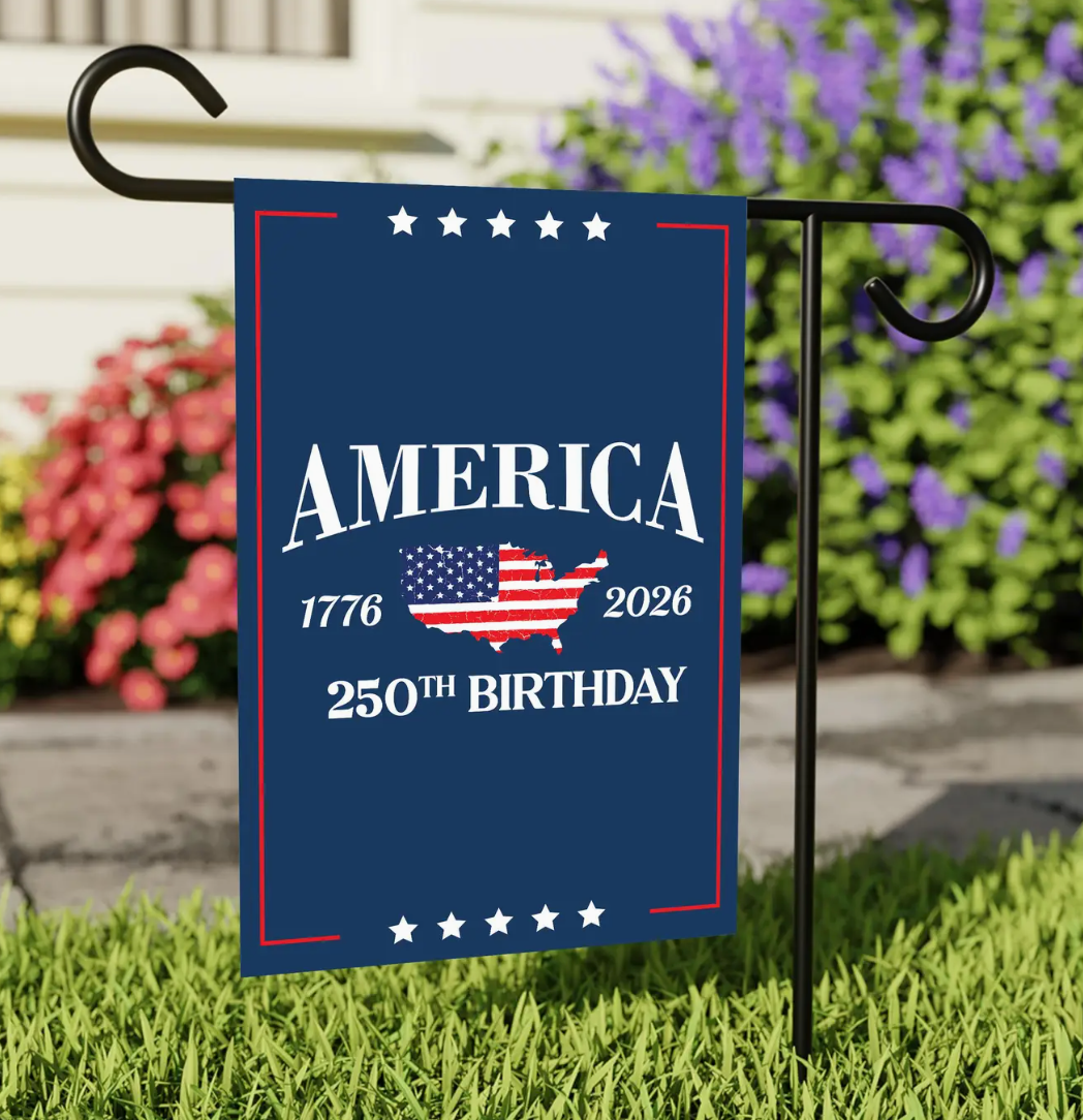Decorative garden flag celebrating America's 250th birthday on a grassy lawn with flowers in the background.