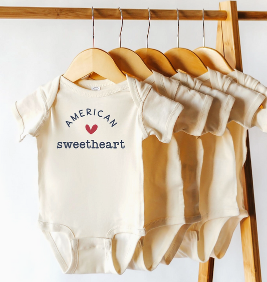 Baby onesie with 'American sweetheart' text on a wooden hanger against a white wall.