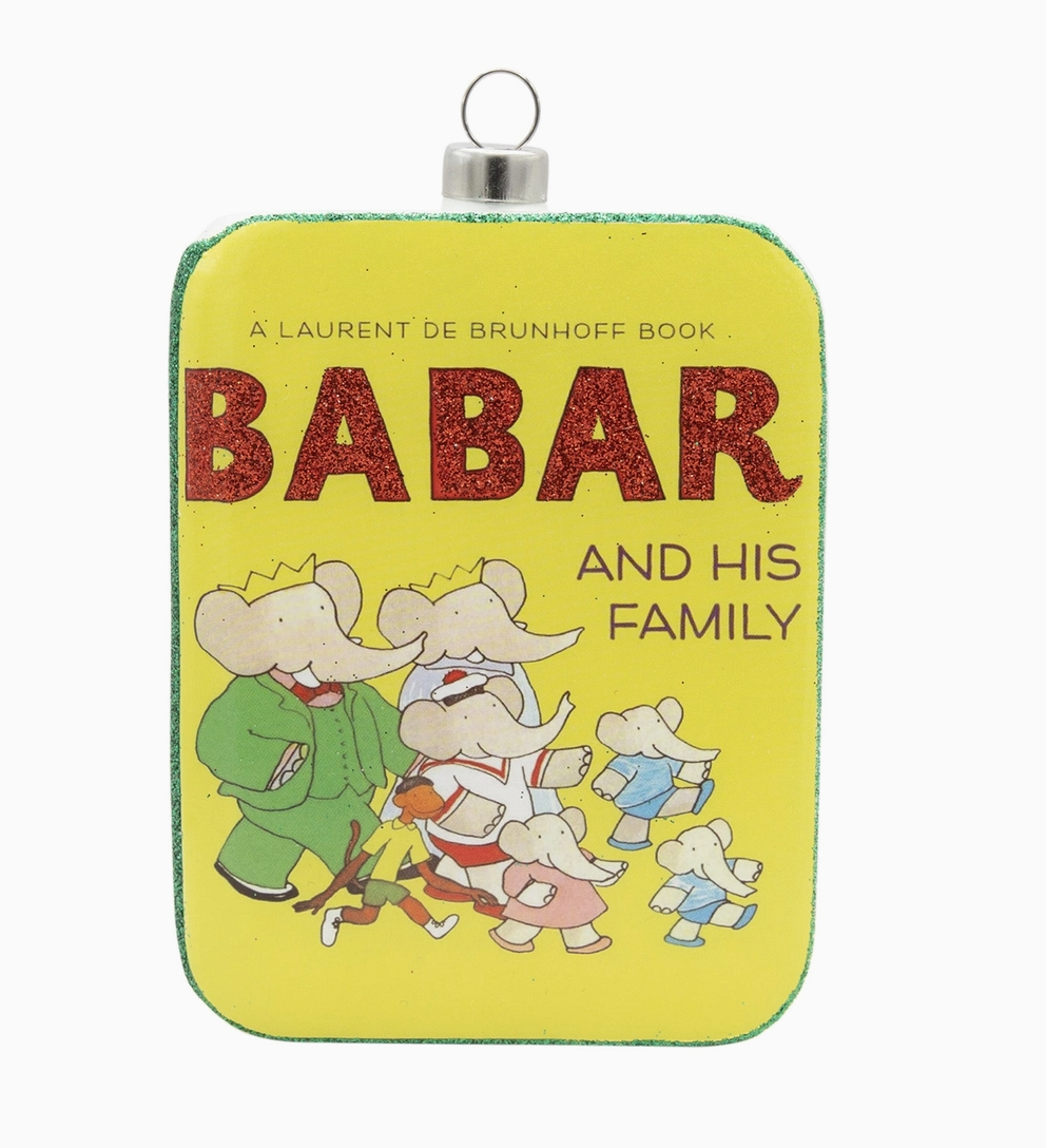 A book ornament shaped like the cover of 'Babar and His Family' by Laurent de Brunhoff. The ornament is bright yellow with a green border, featuring the title in glittery red letters and an illustration of the elephant family.