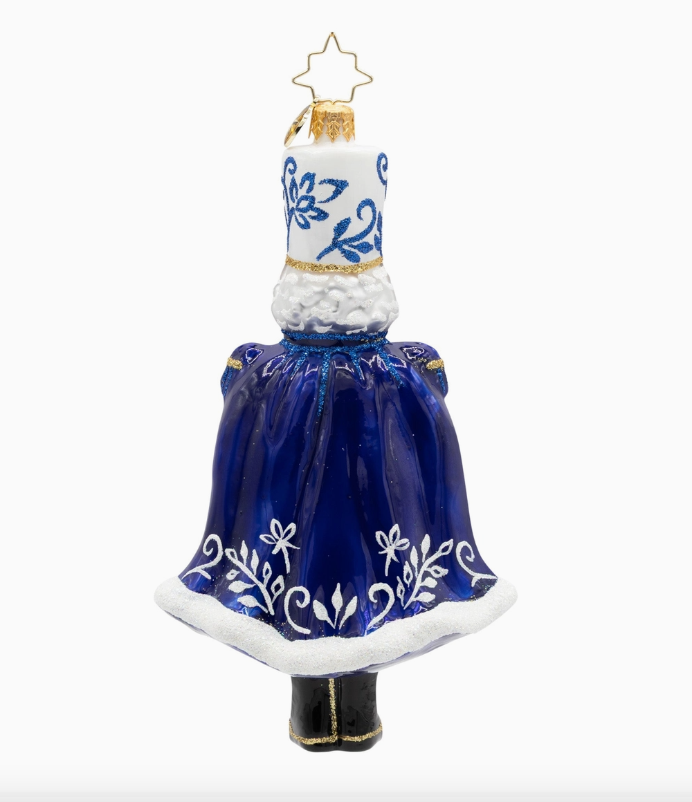 A decorative Christmas ornament shaped like a miniature snowman wearing a blue dress with white floral patterns and a white fluffy collar, topped with a gold star.