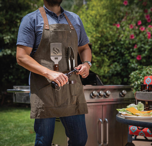 Man in a brown apron standing outdoors by a grill with food and drinks on a table.