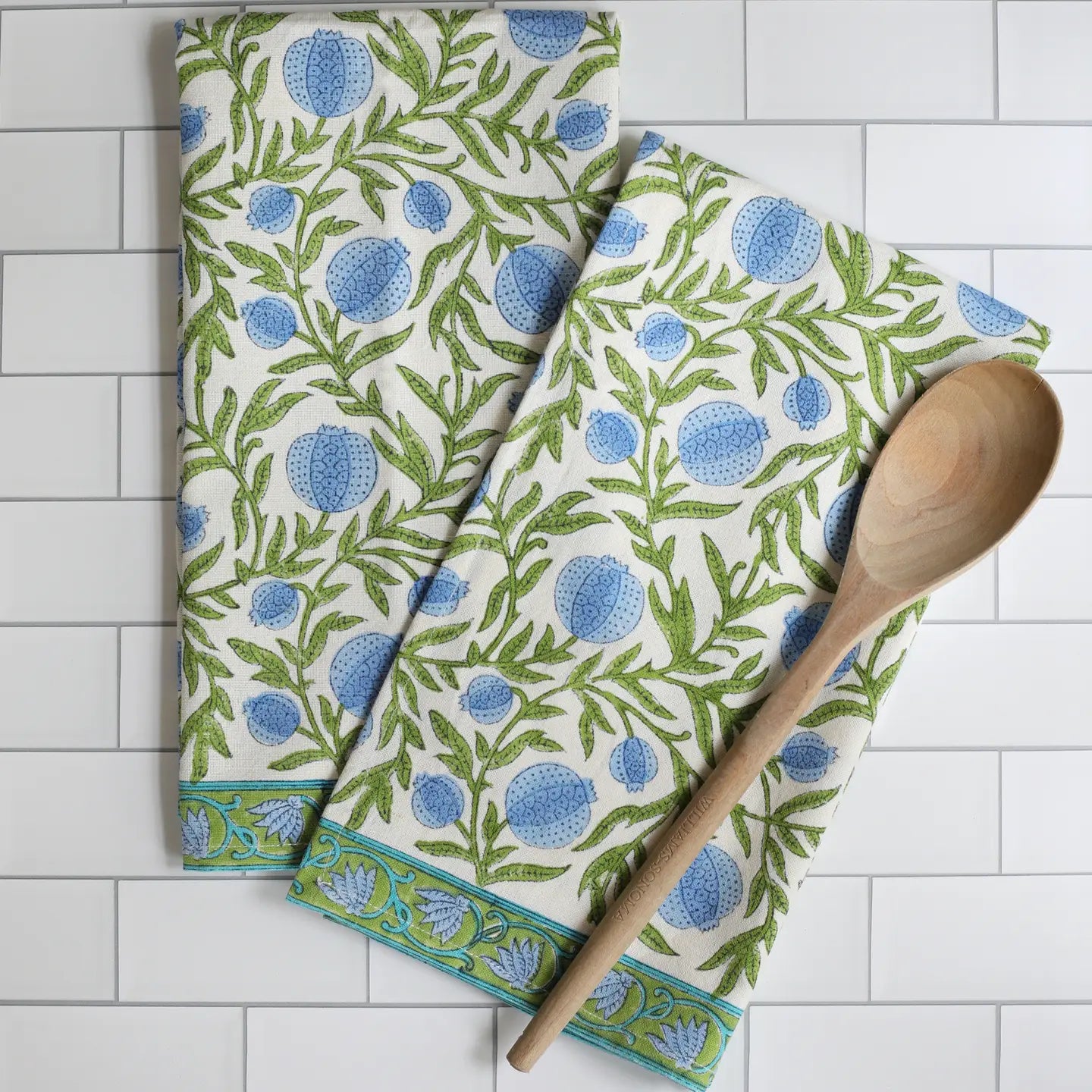 Two patterned towels with blue and green design on a white tiled surface, accompanied by a wooden spoon.