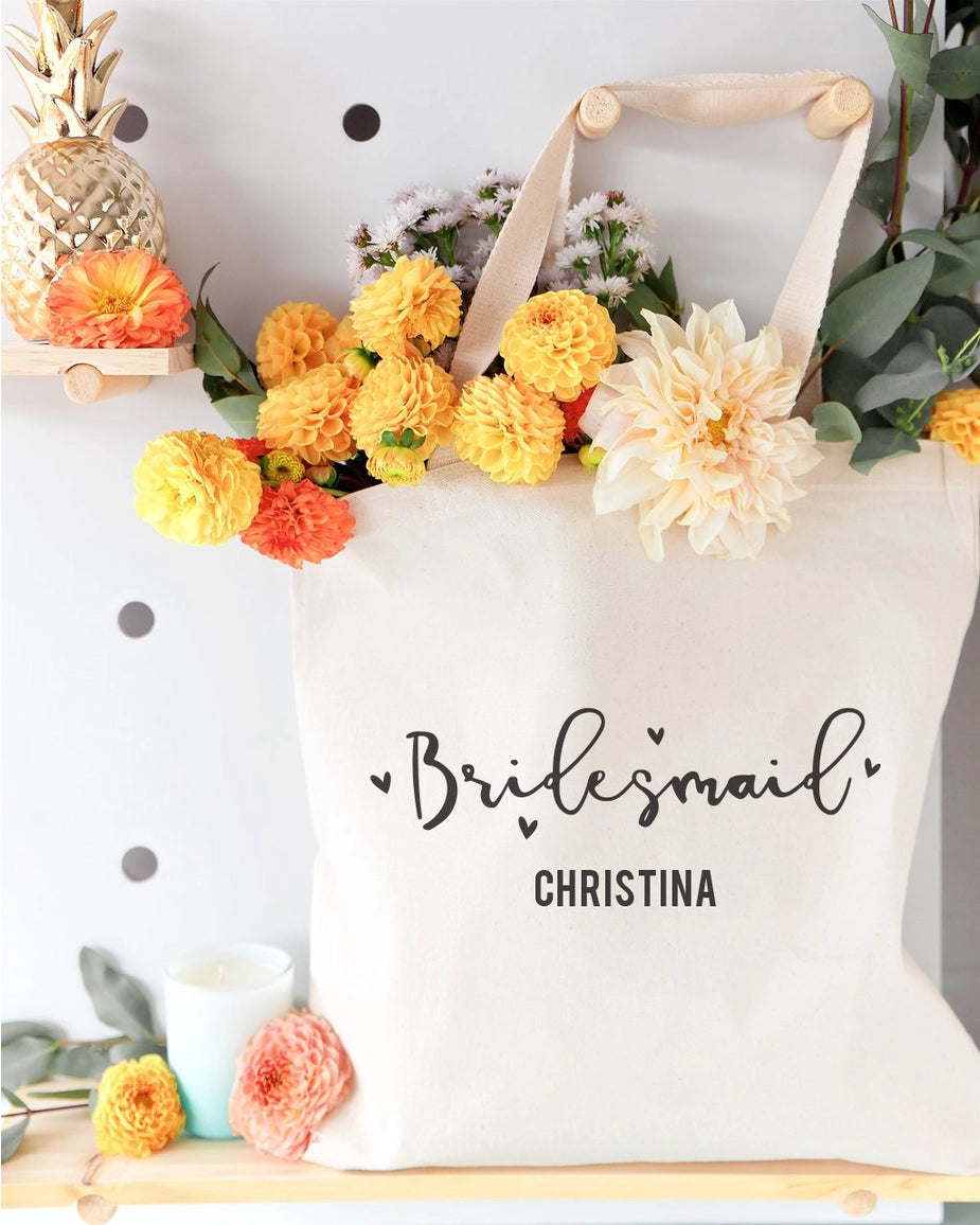 White tote bag with 'Bridesmaid Christina' text, surrounded by flowers and decorative items.