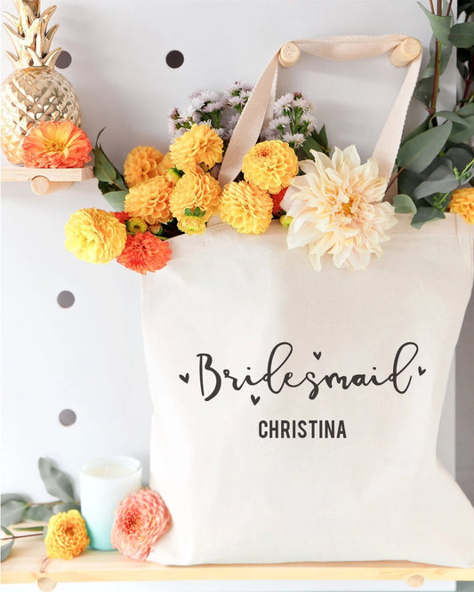 White tote bag with 'Bridesmaid Christina' text, surrounded by flowers and decorative items.