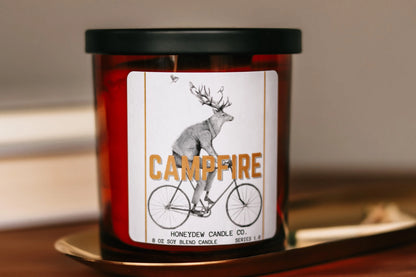 Candle with a label featuring a deer on a bicycle and 'Campfire' text, placed on a surface.