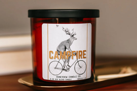 Candle with a label featuring a deer on a bicycle and 'Campfire' text, placed on a surface.