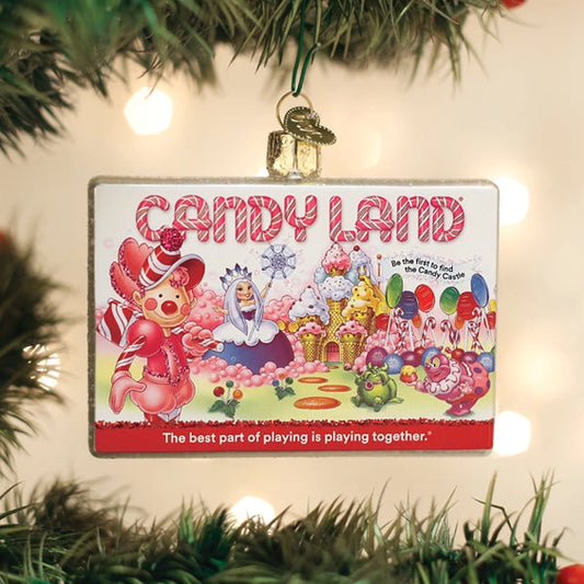 A rectangular Candy Land Christmas ornament hanging from a green tree branch with warm string lights in the background. The ornament features a colorful illustration of characters and candy-themed scenery, with the words 'CANDY LAND' at the top and a red slogan at the bottom.