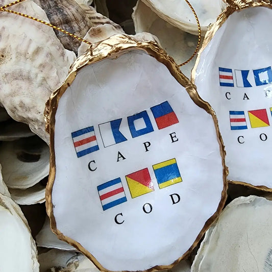 A white oyster shell ornament with a gold-rimmed edge, featuring printed nautical signal flags and the words 'CAPE COD' on the front, displayed among other shells.