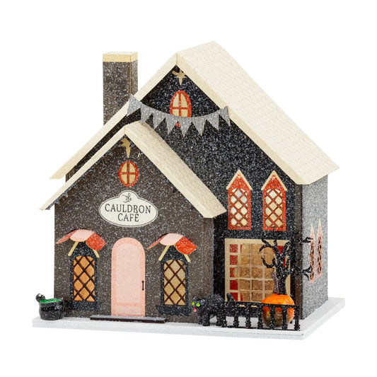 A decorative miniature house-shaped gingerbread cafe ornament labeled 'The Cauldron Cafe' on a white background. The house is dark brown with glittery texture, a cream-colored roof, orange window accents, a pink door, and Halloween-themed decorations including a small black cat, pumpkin, and tree.