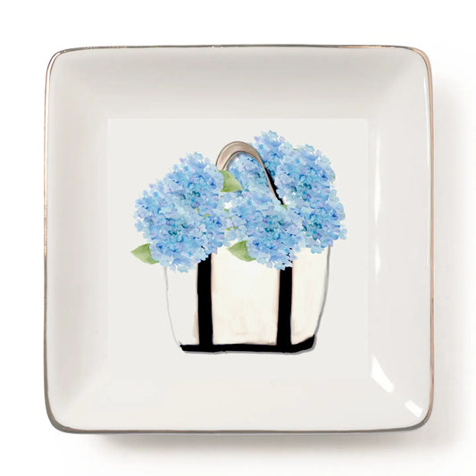 A square white ceramic plate with rounded corners and a thin metallic gold rim, featuring a central illustration of light blue hydrangea flowers in a white basket with black handles.