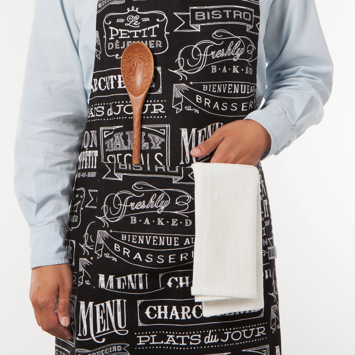 A person wearing a black apron with a white vintage bistro and bakery print, holding a wooden spoon and a white towel in front of a light blue shirt.