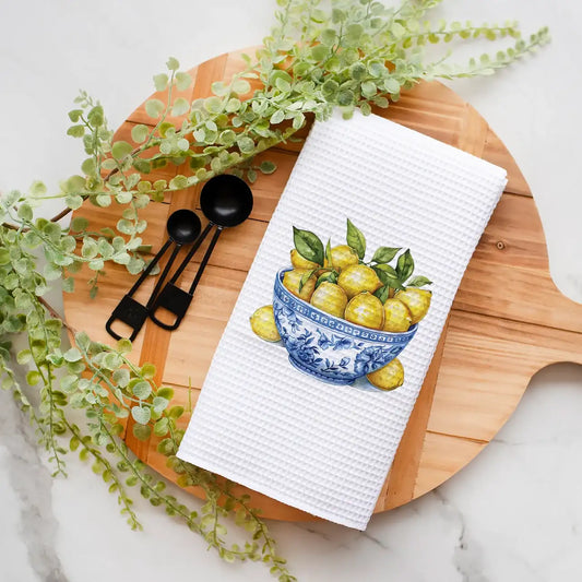 White towel with a lemon design on a wooden cutting board with black spoons and greenery.
