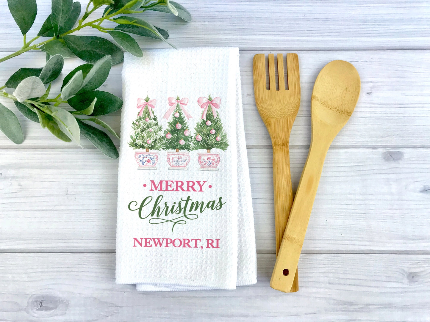 A white waffle-weave kitchen towel with a Christmas tree print and 'MERRY Christmas Newport, RI' text, styled on a light wood surface with a bamboo spatula and spoon and green leaves.