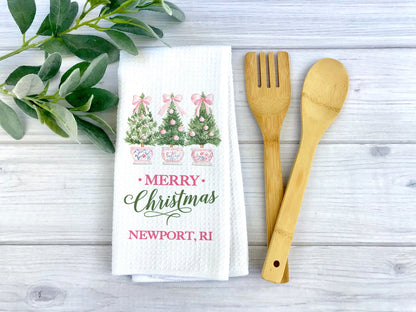 A white waffle-weave kitchen towel with a Christmas tree print and 'MERRY Christmas Newport, RI' text, styled on a light wood surface with a bamboo spatula and spoon and green leaves.